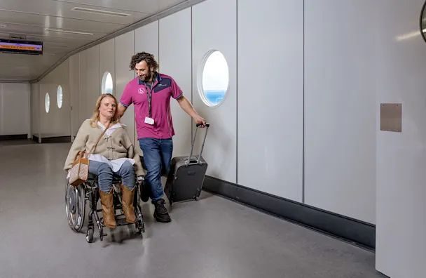 Airport staff member assisting elderly passenger in wheelchair through terminal corridor