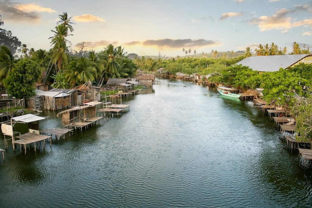 Stilt houses line a calm river channel through Amazon rainforest with distant city skyline