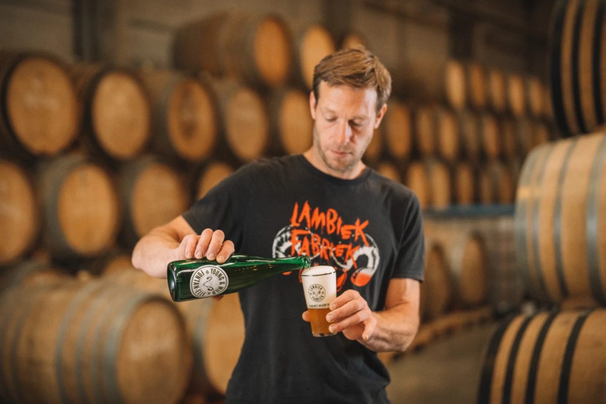 Brewer holding a bottle and glass of lambic beer surrounded by oak barrels.