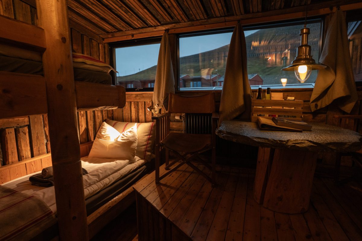 Cozy wooden cabin interior with bed and desk overlooking Arctic landscape at dusk