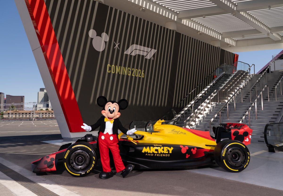 Mickey Mouse stands beside a yellow and red Formula 1 car at a modern stadium venue