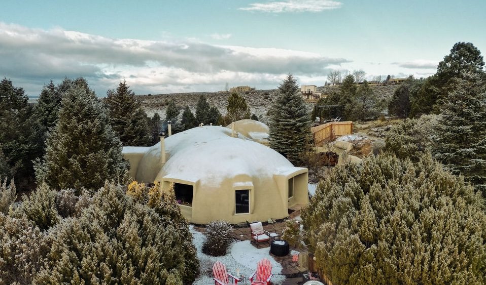 Dome-shaped yellow rental home surrounded by evergreen trees with mountains in background