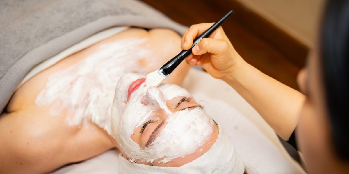 Esthetician applying white facial mask treatment to client's face with brush