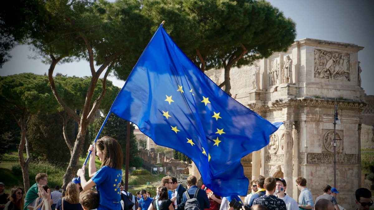 EU flag waving at a historic European landmark with tourists gathered nearby