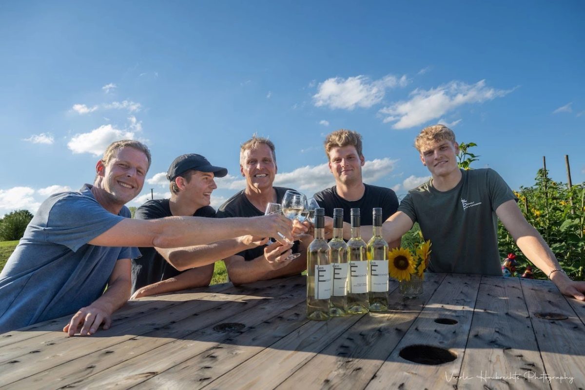 Five people enjoying wine tasting at a vineyard with bottles displayed on a wooden table