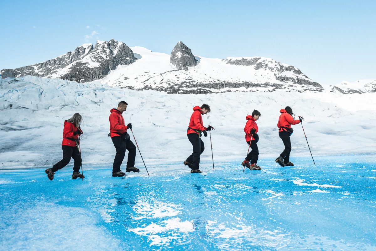 Group of hikers in red jackets crossing bright blue glacial ice with snow-capped mountains in background