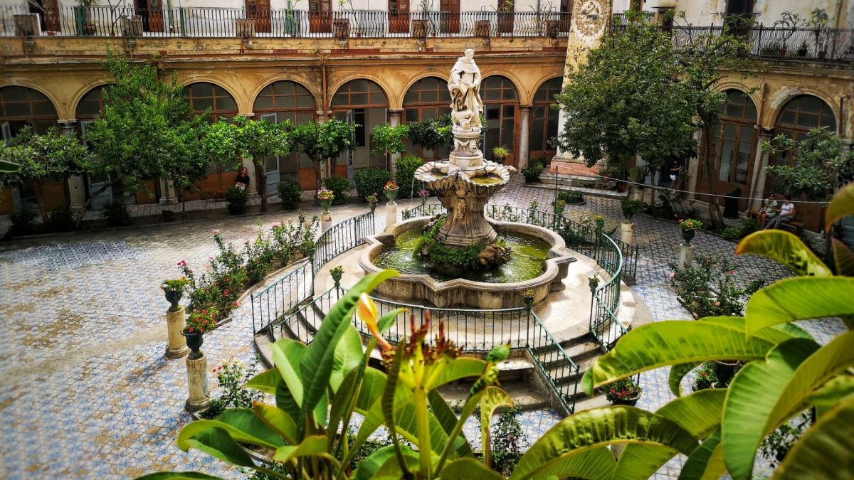 Historic courtyard in Palermo with arched colonnade, central fountain, and lush Mediterranean plants