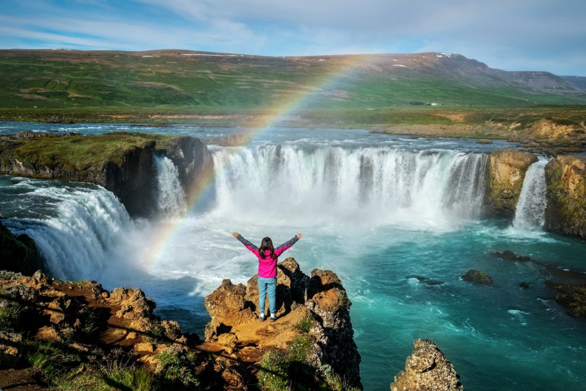Person in pink jacket with arms raised on rocky outcrop overlooking Godafoss waterfall in Iceland