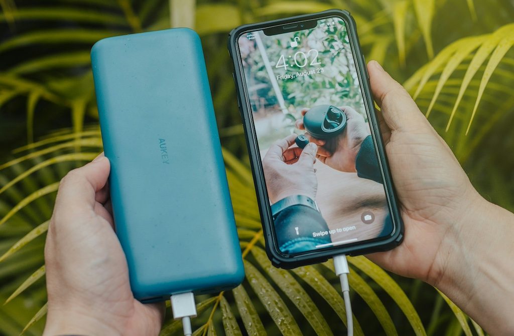 Hands holding a smartphone and blue power bank against a leafy green background