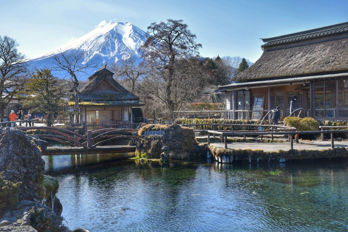 Traditional Japanese buildings beside a clear pond with Mount Fuji snow-capped in the background
