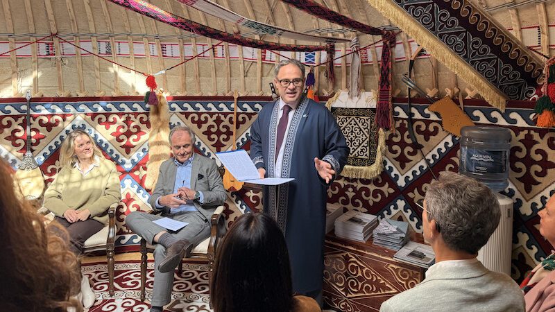 Man in blue suit speaking to seated guests in traditional Kazakh yurt with ornate carpets