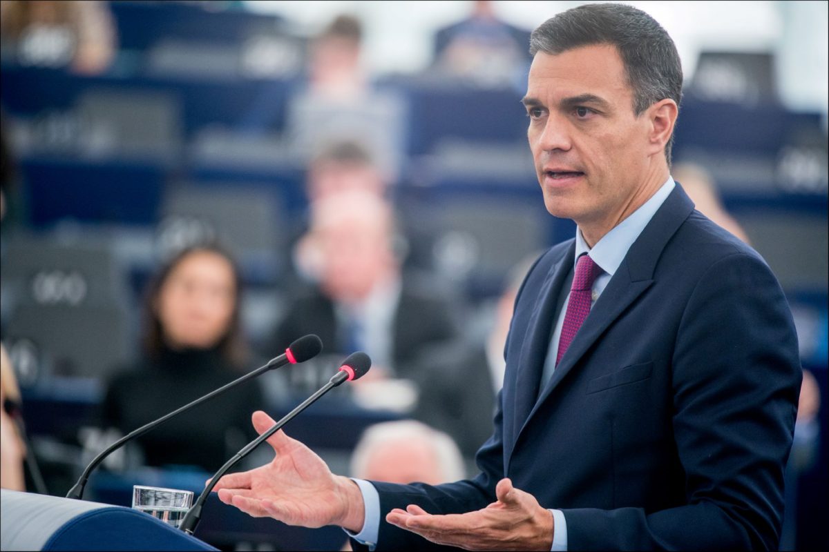 Man in dark suit and purple tie speaking at podium with microphone in parliament