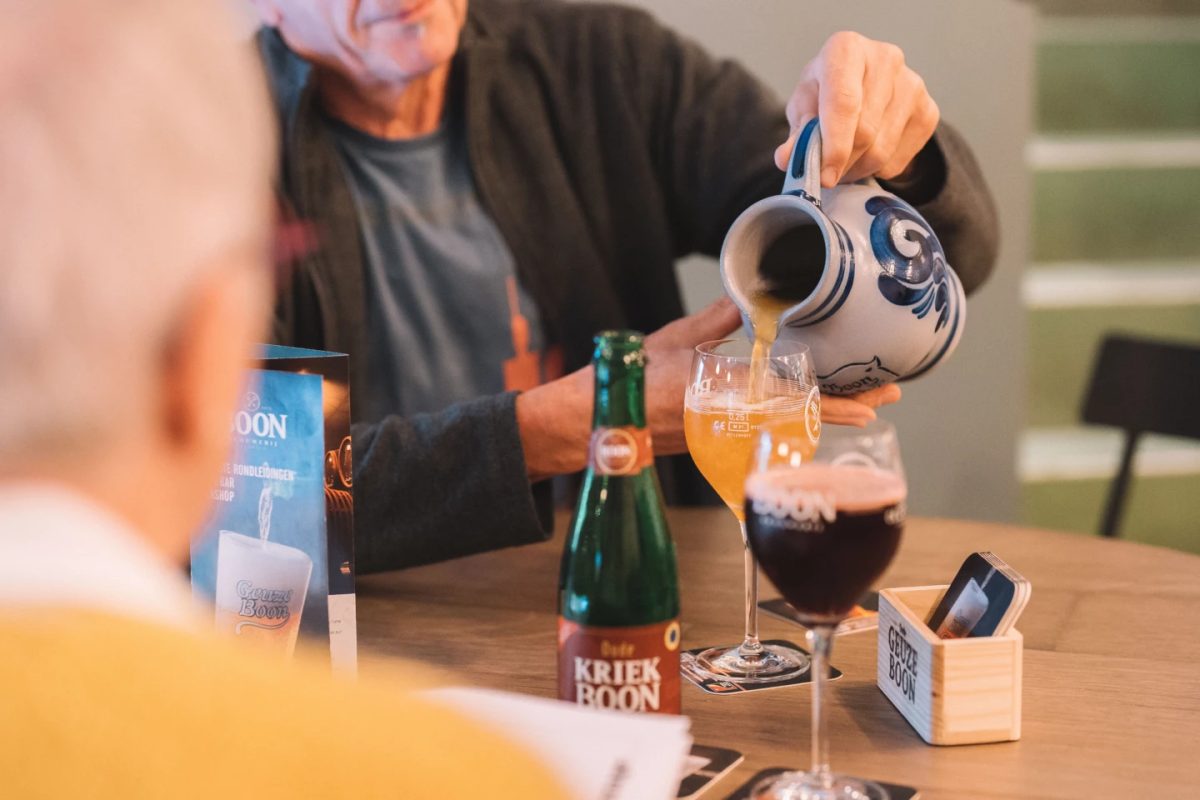 Man pouring lambic beer into a glass, with bottles and tasting notes on a wooden table.