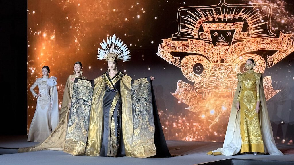 Models wearing elaborate gold and cream traditional Chinese costumes on a runway with dramatic lighting