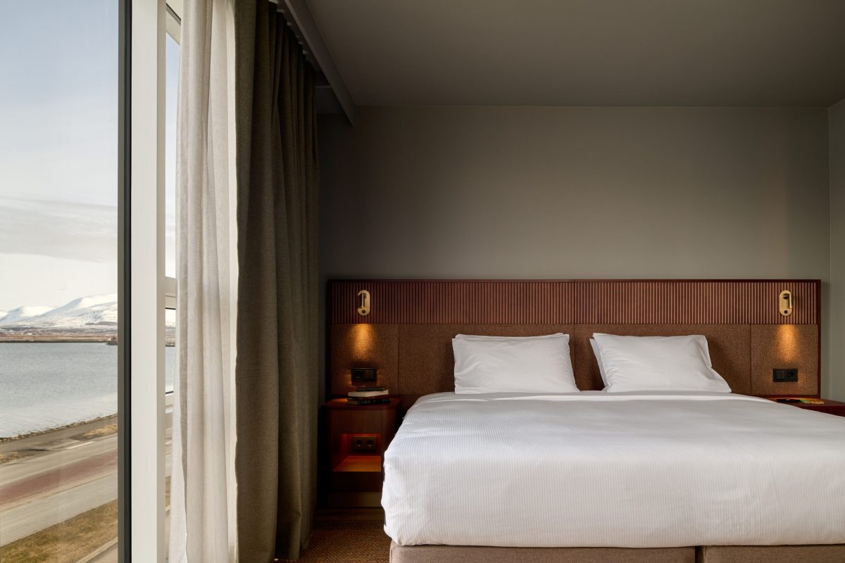 Modern hotel bedroom with white bedding, wooden headboard, and panoramic window overlooking Iceland's coastal landscape