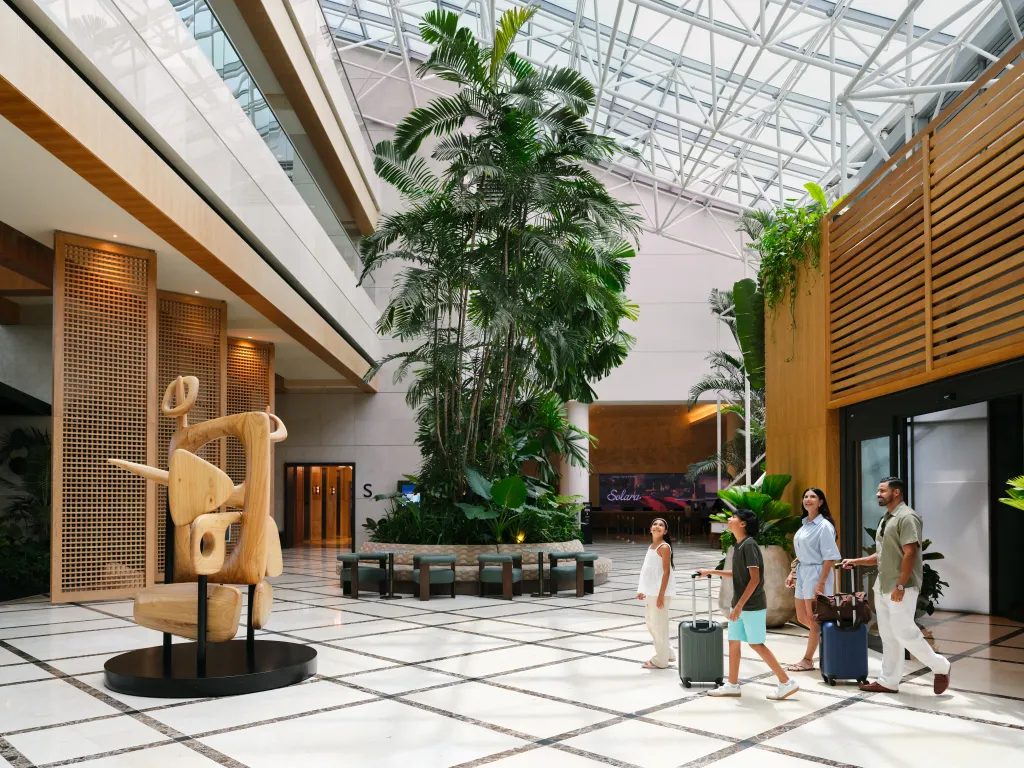 Modern hotel lobby with lush greenery, a sculpture, and a family arriving with luggage.