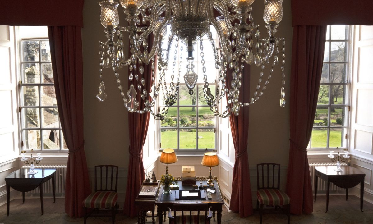 Ornate room with burgundy curtains, crystal chandelier, and tall windows overlooking gardens