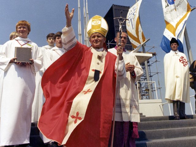 Religious ceremony with clergy in ceremonial robes and flags on what appears to be a ship or vessel
