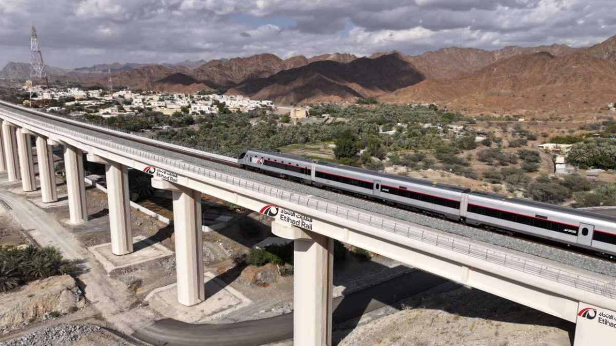 Modern elevated train on concrete viaduct with desert mountains and green oasis city in background