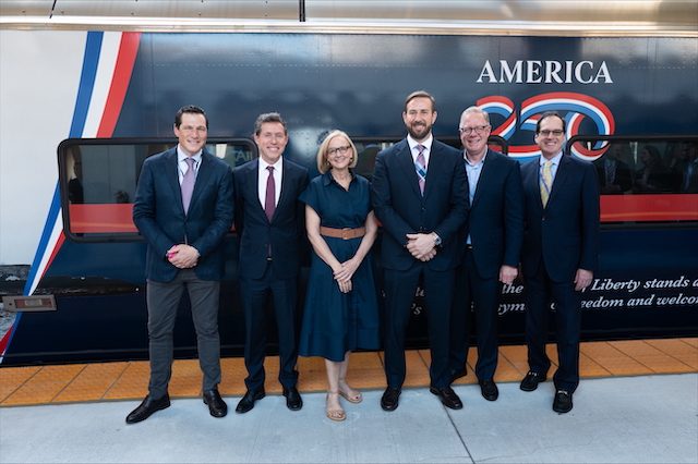 Six officials stand in front of Brightline's patriotic Freedom Express train decorated with American flag colors