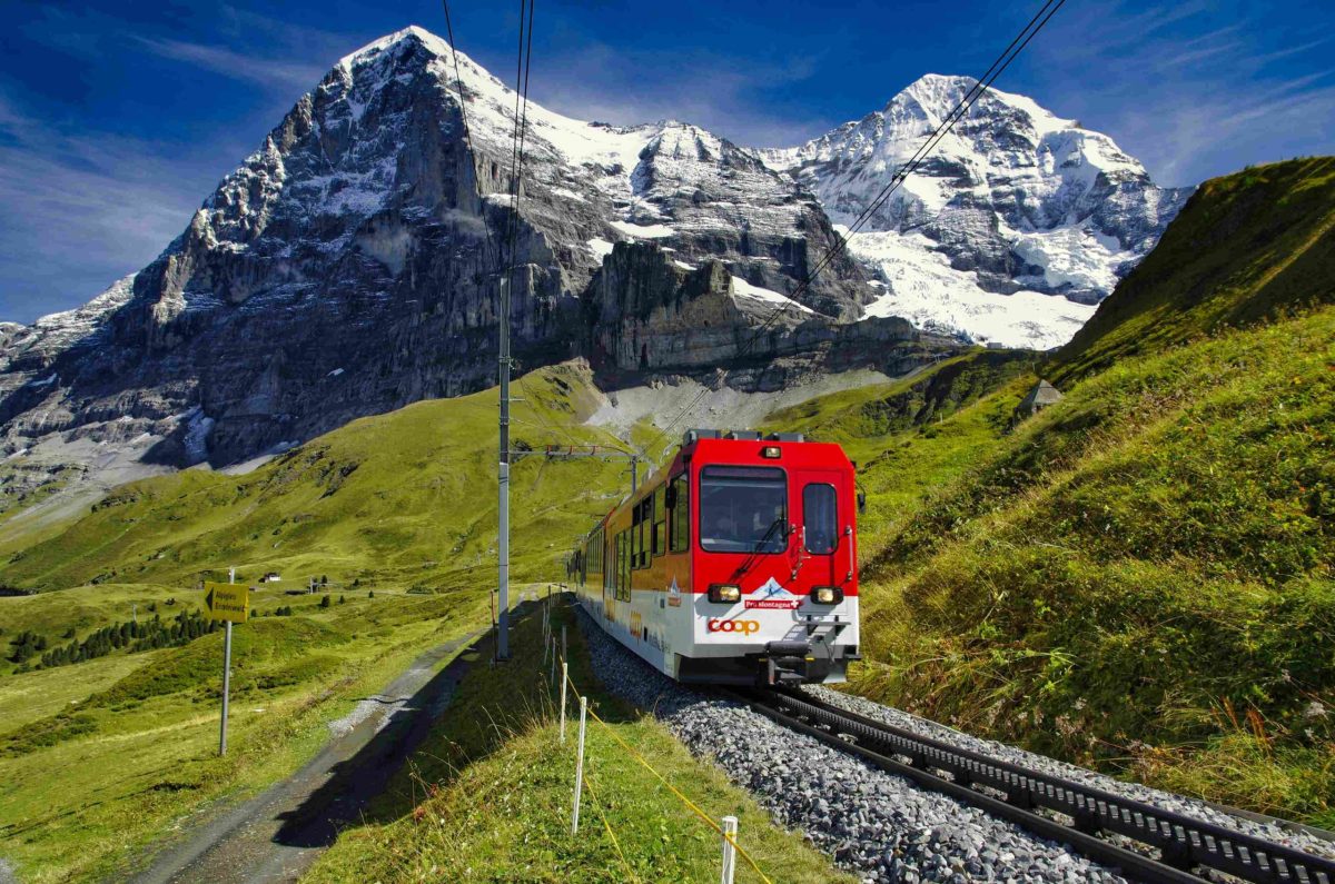Red and yellow Swiss train winding through a green valley with snow-capped mountains in the background.