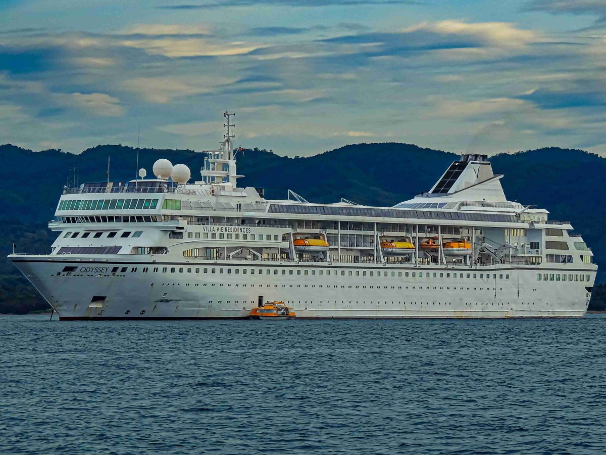 Large cruise ship docked against a backdrop of lush green hills and a cloudy sky.
