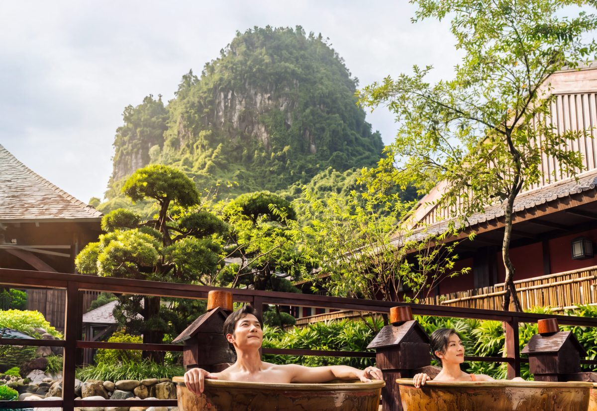 Two people relax in outdoor stone tubs overlooking a lush, mountainous landscape.