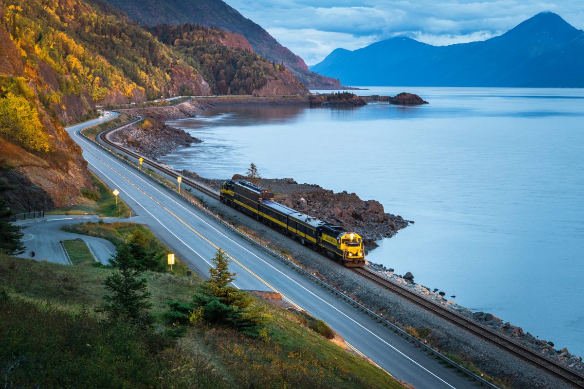 Train travels along a scenic coastal route with mountains and water.