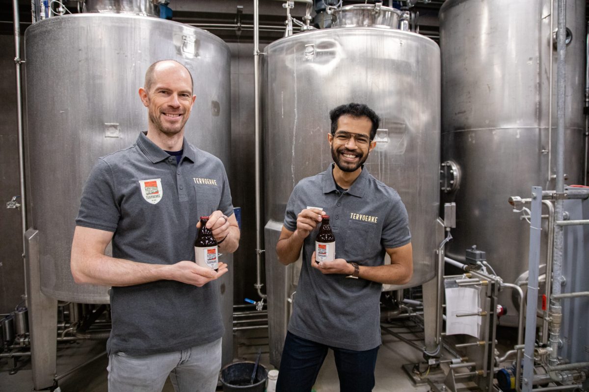 Two brewers holding beer glasses in front of large stainless steel fermentation tanks