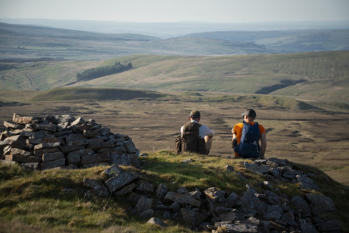Two hikers sitting on rocky outcrop overlooking rolling Cumbrian valleys and moorland