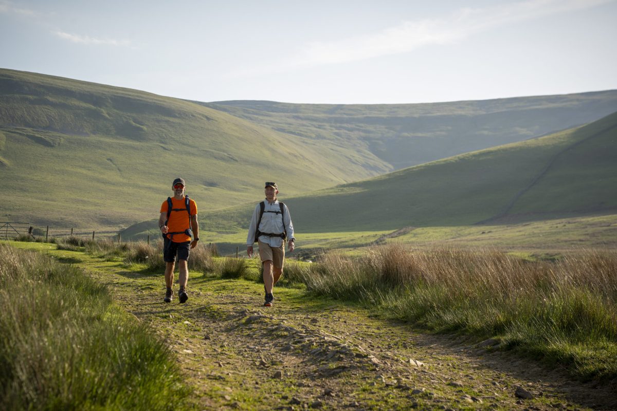 Two hikers walking on a dirt trail through rolling green hills and valleys