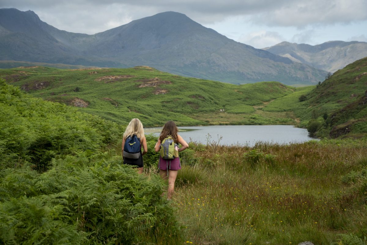 Two hikers with backpacks walking through green moorland toward a mountain lake