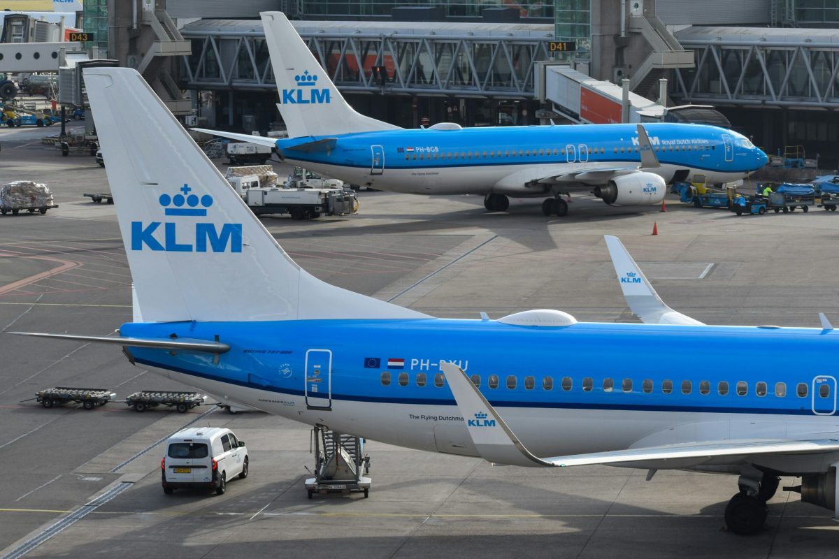 Two blue and white KLM aircraft parked at Amsterdam Airport tarmac with ground vehicles nearby