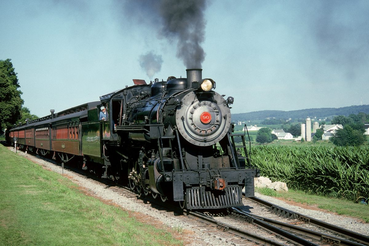 Vintage steam train pulls passenger cars through a rural landscape.