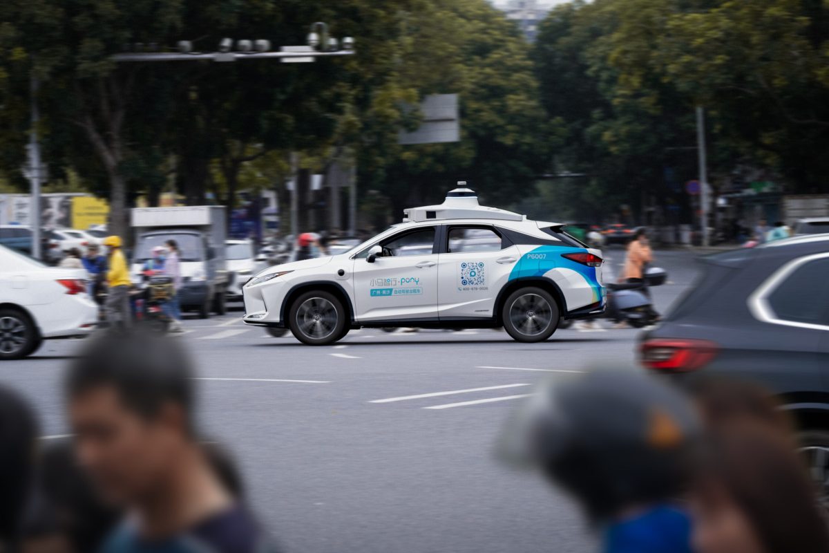 White and blue autonomous vehicle on city street surrounded by pedestrians and other traffic