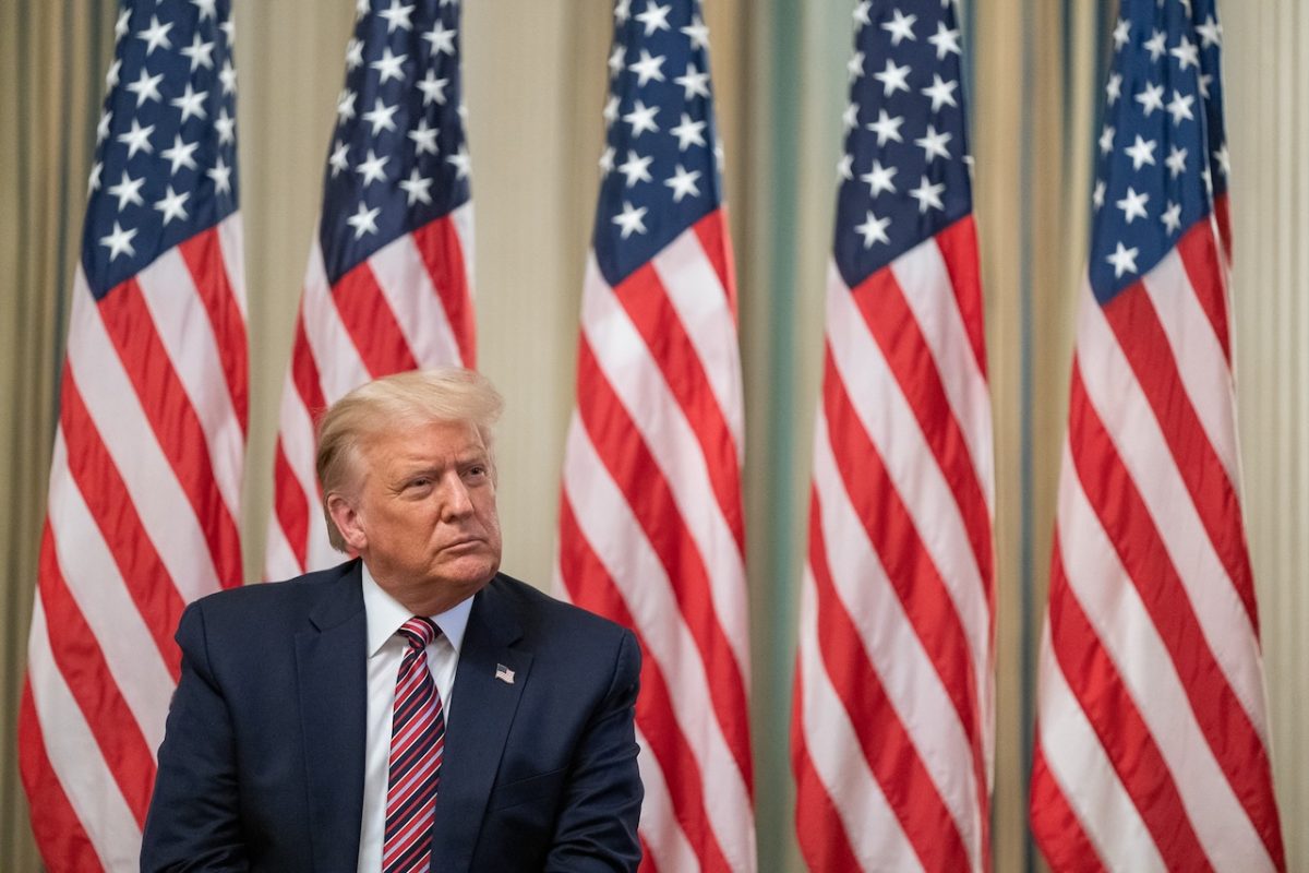 Man in dark suit and red tie seated in front of five American flags